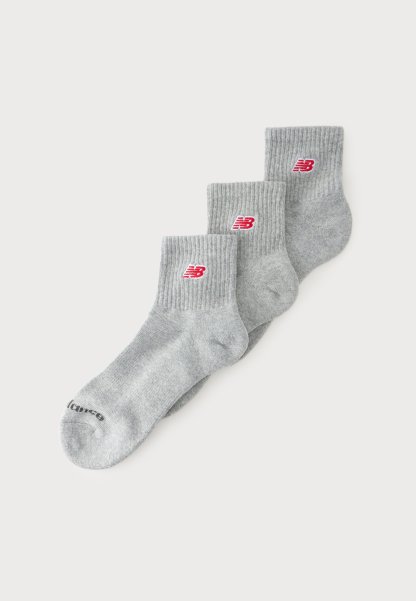 Tres pares de calcetines tobilleros grises con puños acanalados y logotipos rojos de New Balance están apilados sobre un fondo blanco.