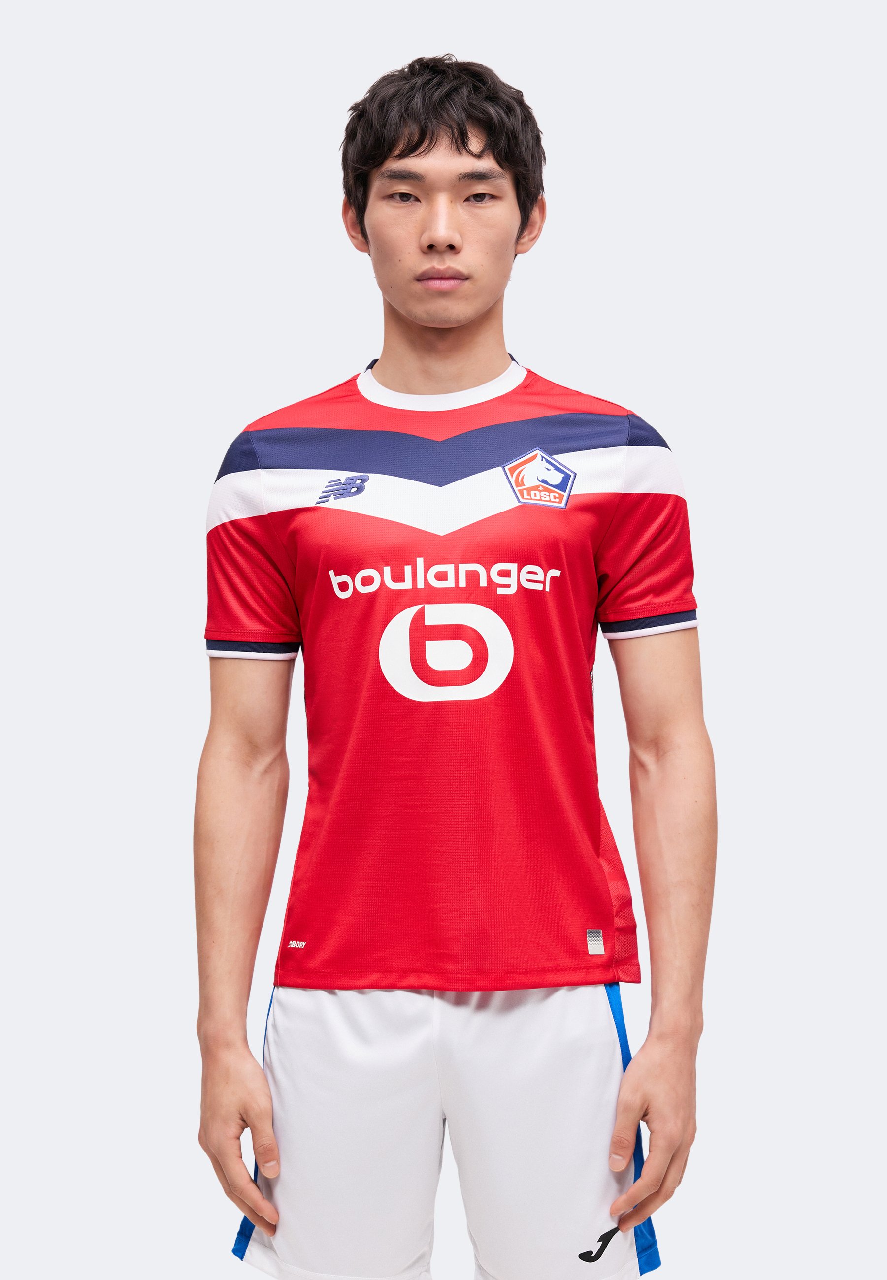 Joven hombre está de pie mirando de frente, vistiendo una camiseta de fútbol LOSC Lille en rojo, blanco y azul, y pantalones cortos blancos sobre un fondo simple.