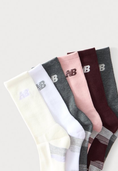 Seis pares de calcetines New Balance tipo crew en blanco, gris, rosa, granate y gris oscuro, dispuestos superpuestos sobre un fondo claro.