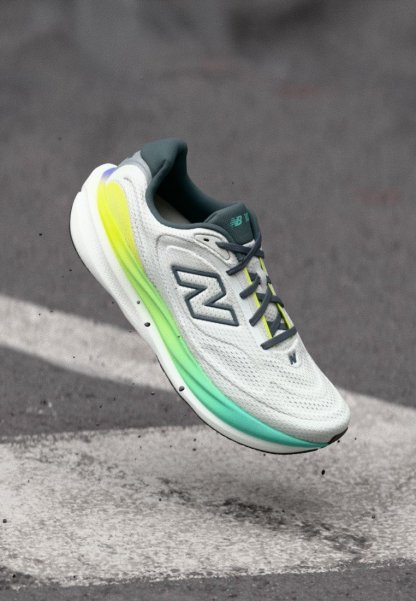 Zapatilla de correr New Balance blanca y gris con detalles en neón amarillo y verde suspendida en el aire sobre el pavimento.