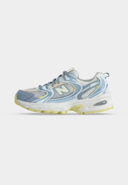 Una única zapatilla de correr New Balance azul claro y blanca con detalles amarillos en la suela y detalles de tela de malla sobre un fondo liso.