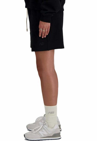 Persona que lleva shorts negros, zapatillas blancas New Balance y calcetines crema hasta la pantorrilla con el logo de New Balance, de pie de lado sobre fondo blanco.
