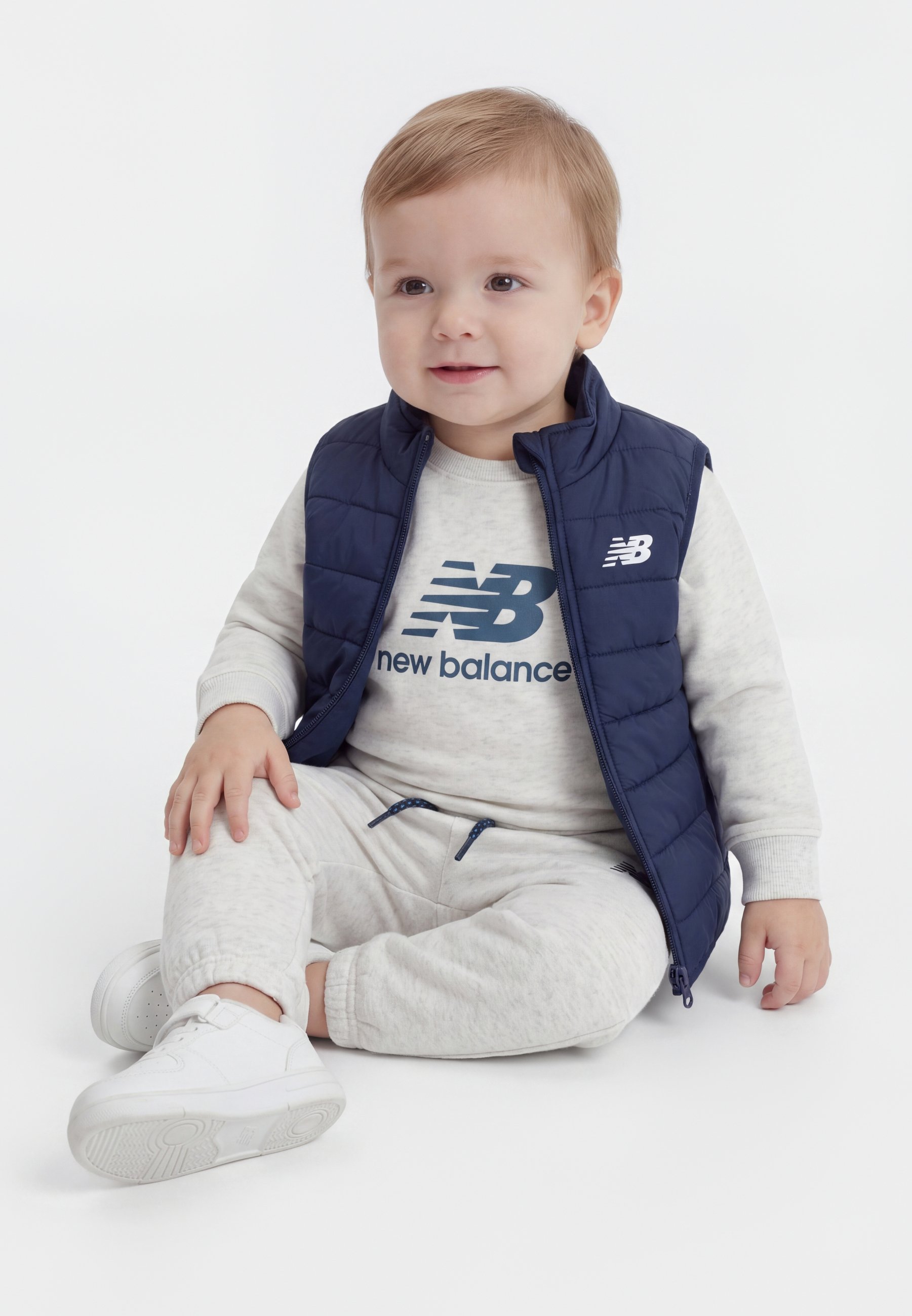 Niño pequeño con cabello castaño claro sentado, vestido con un chaleco azul marino acolchado, sudadera y pantalones gris claro de New Balance, y zapatillas blancas.