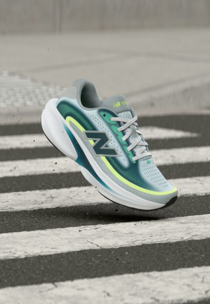 Zapatilla de running New Balance en azul claro y verde con suela blanca, mostrada en el aire sobre un paso de peatones de cebra en un pavimento de asfalto.
