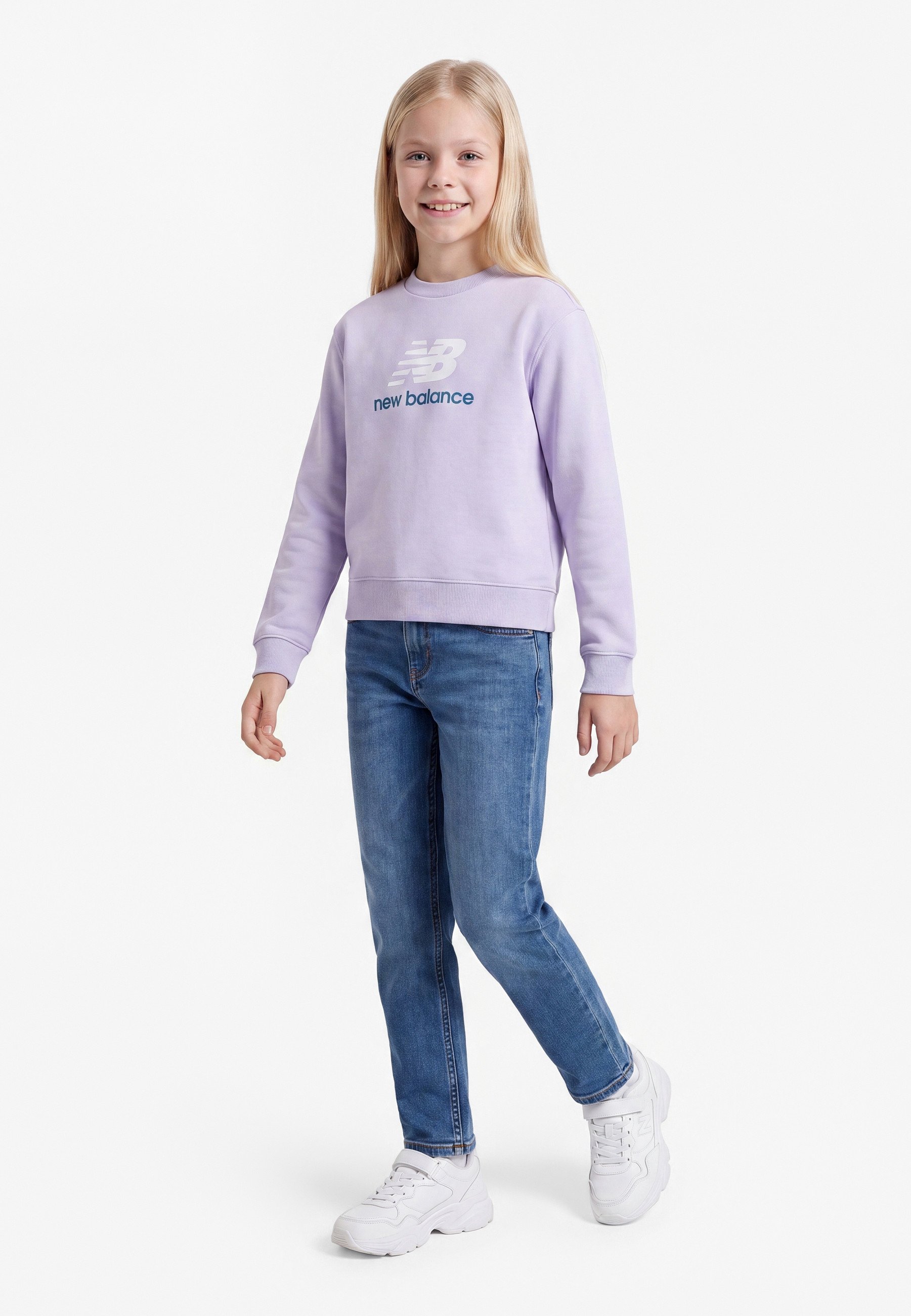 Niña sonriente de pie, con sudadera New Balance de color púrpura claro, vaqueros azules y zapatillas blancas sobre un fondo blanco liso.