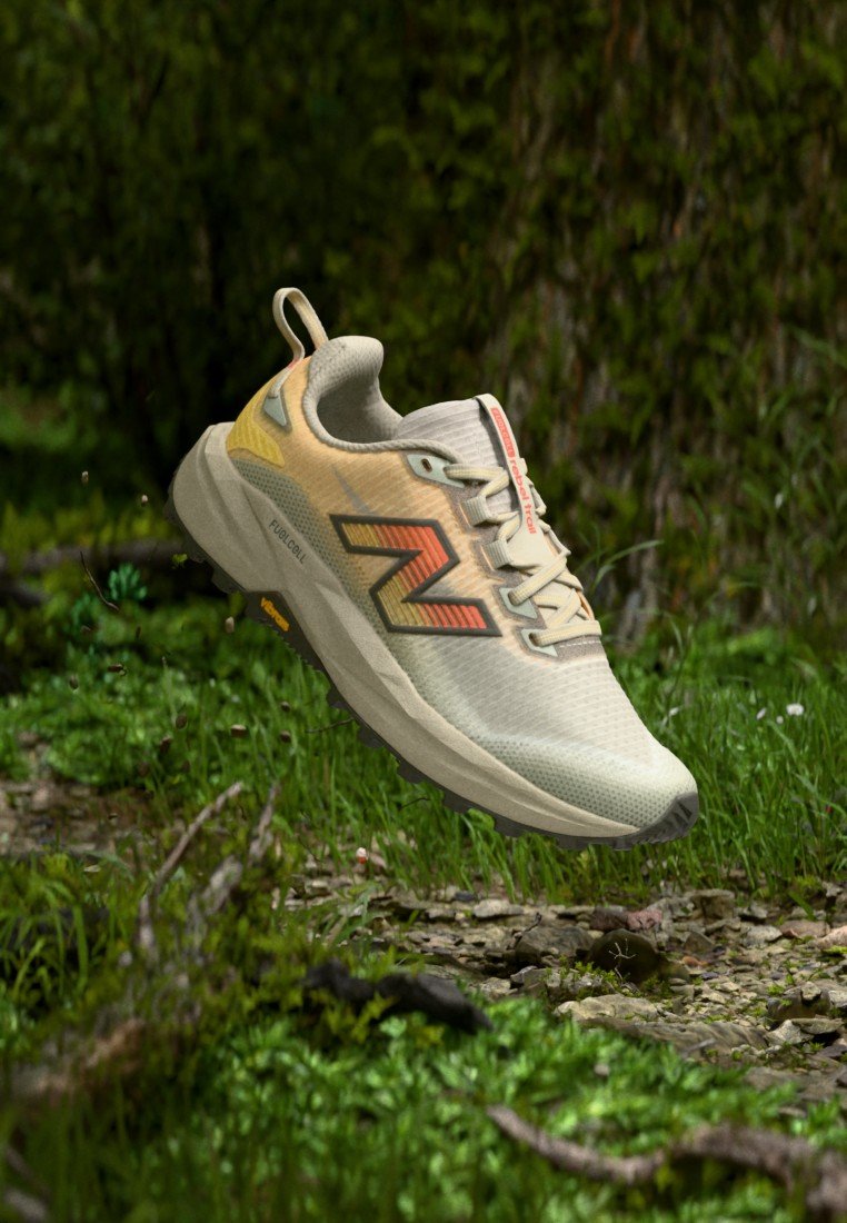 Zapatilla de correr beige claro y amarilla con el logo "N" naranja, suspendida en el aire sobre un suelo de bosque con musgo verde y pequeñas piedras.