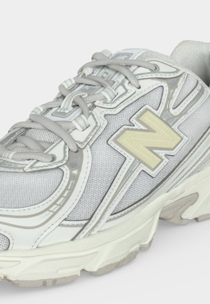 Zapatilla deportiva blanca y gris con tejido de malla, cierre con cordones y gran logo "N" en el lateral.