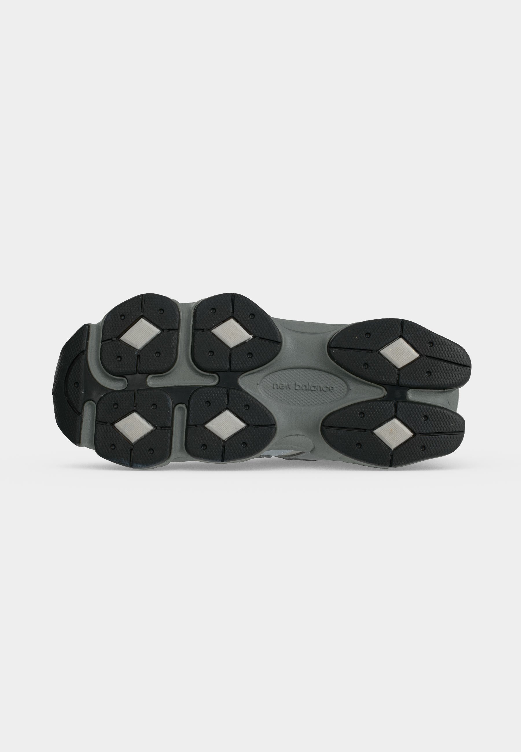 Suela de zapato New Balance gris y negro con dibujos en la banda de rodadura y zonas de agarre blancas en forma de diamante sobre fondo blanco.