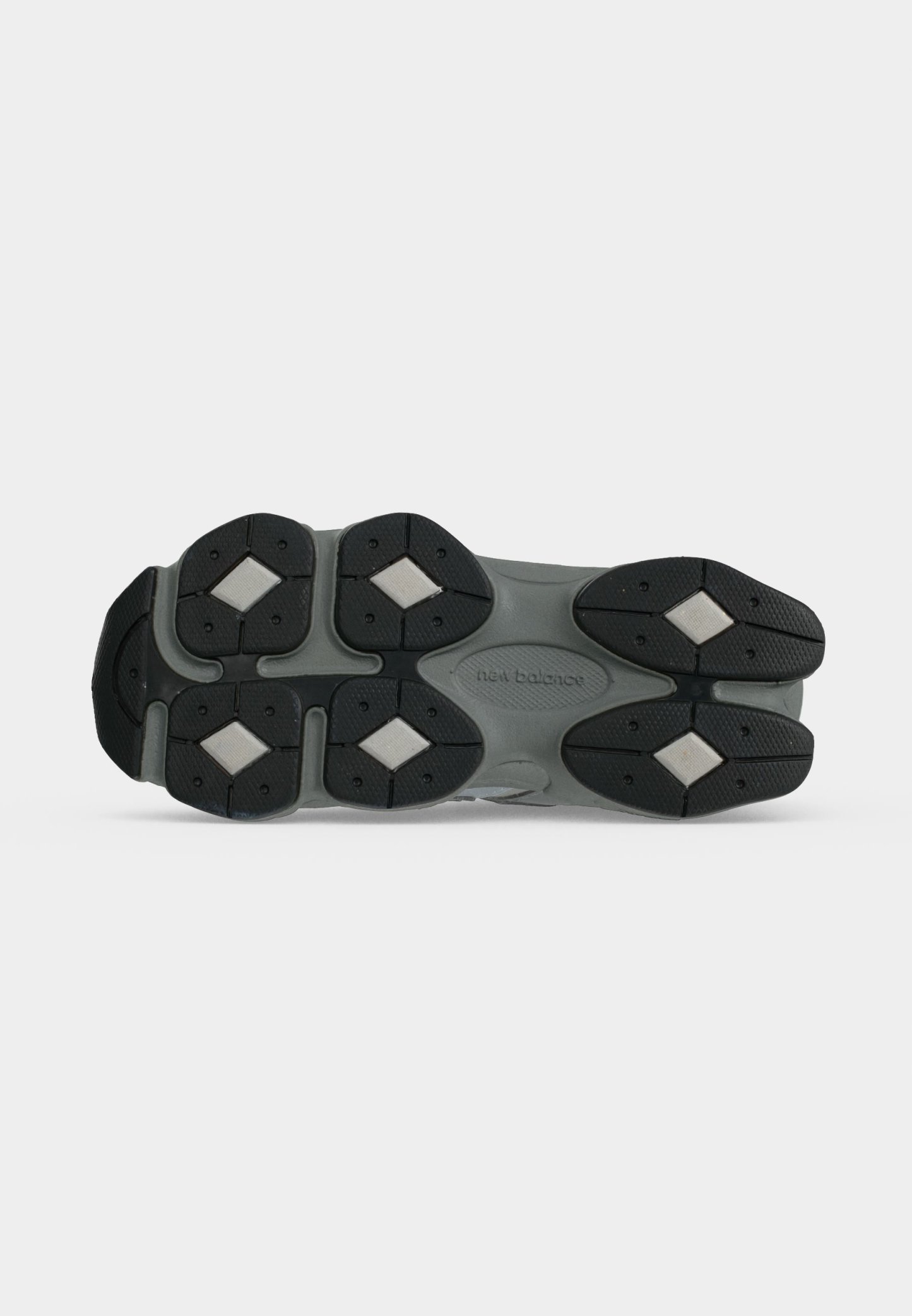Suela de zapato New Balance gris y negro con dibujos en la banda de rodadura y zonas de agarre blancas en forma de diamante sobre fondo blanco.