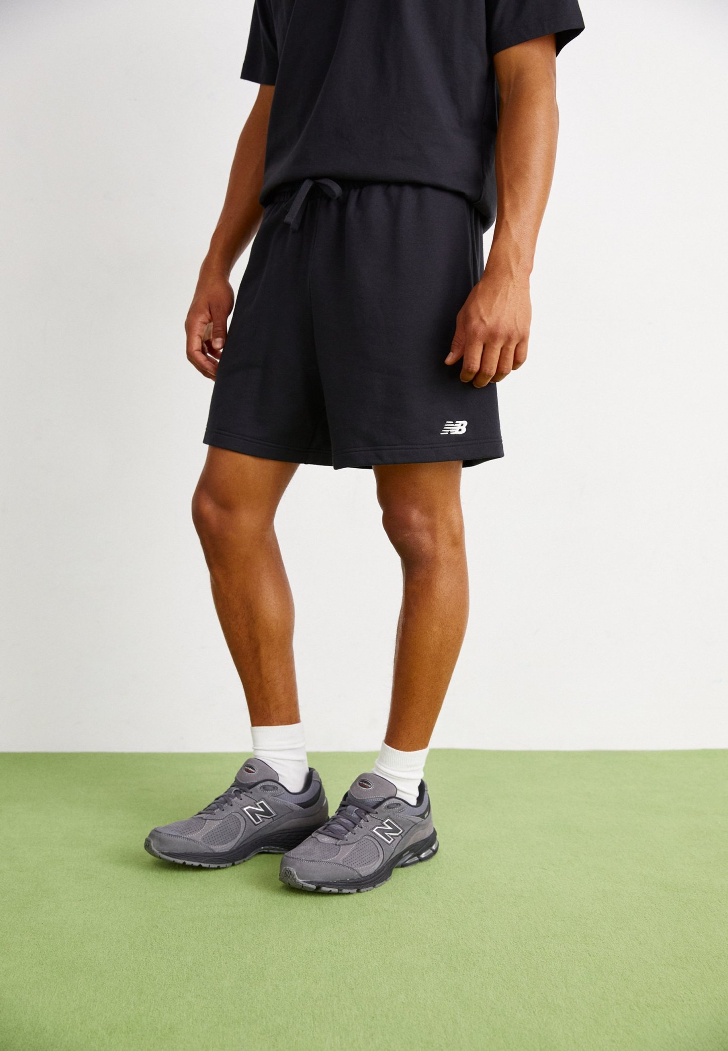 Pantalones cortos de algodón negros con una cinturilla elástica, combinados con zapatillas New Balance grises que presentan detalles de malla y goma, sobre un suelo verde.