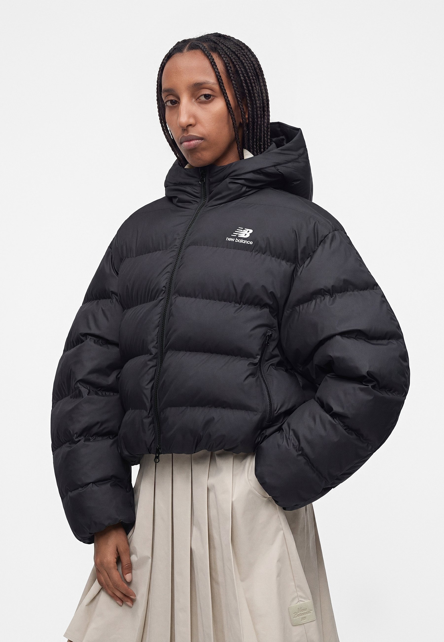 Mujer con cabello trenzado que lleva una chaqueta acolchada negra de New Balance y una falda plisada beige claro, de pie frente a un fondo blanco liso.
