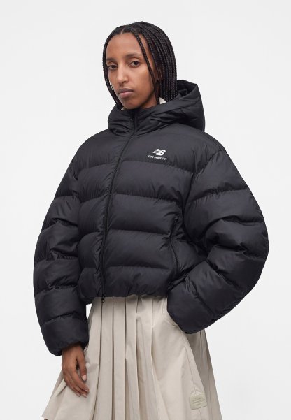 Mujer con cabello trenzado que lleva una chaqueta acolchada negra de New Balance y una falda plisada beige claro, de pie frente a un fondo blanco liso.