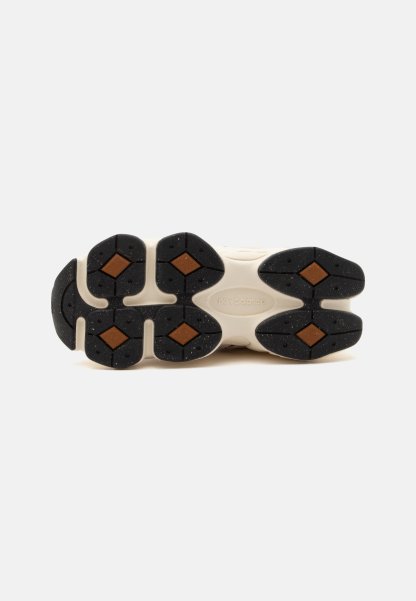 Suela de una zapatilla con tacos de goma segmentados en negro y marrón sobre una base beige, mostrando el nombre de la marca en relieve en el centro.