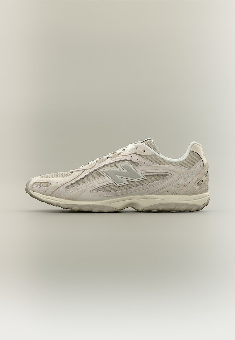 Zapatilla New Balance de color gris claro con parte superior de malla y ante, diseño con cordones y suela de goma texturizada, vista lateral sobre fondo neutro.
