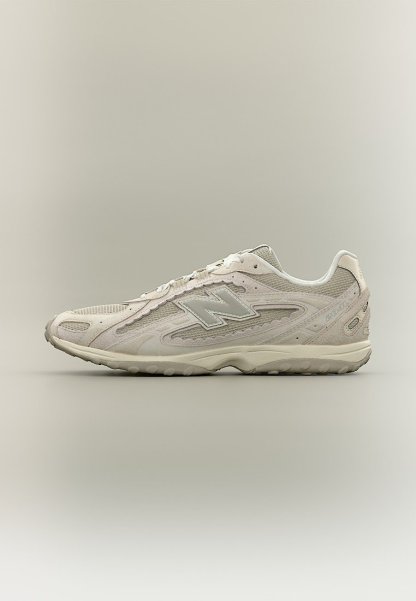 Zapatilla New Balance de color gris claro con parte superior de malla y ante, diseño con cordones y suela de goma texturizada, vista lateral sobre fondo neutro.
