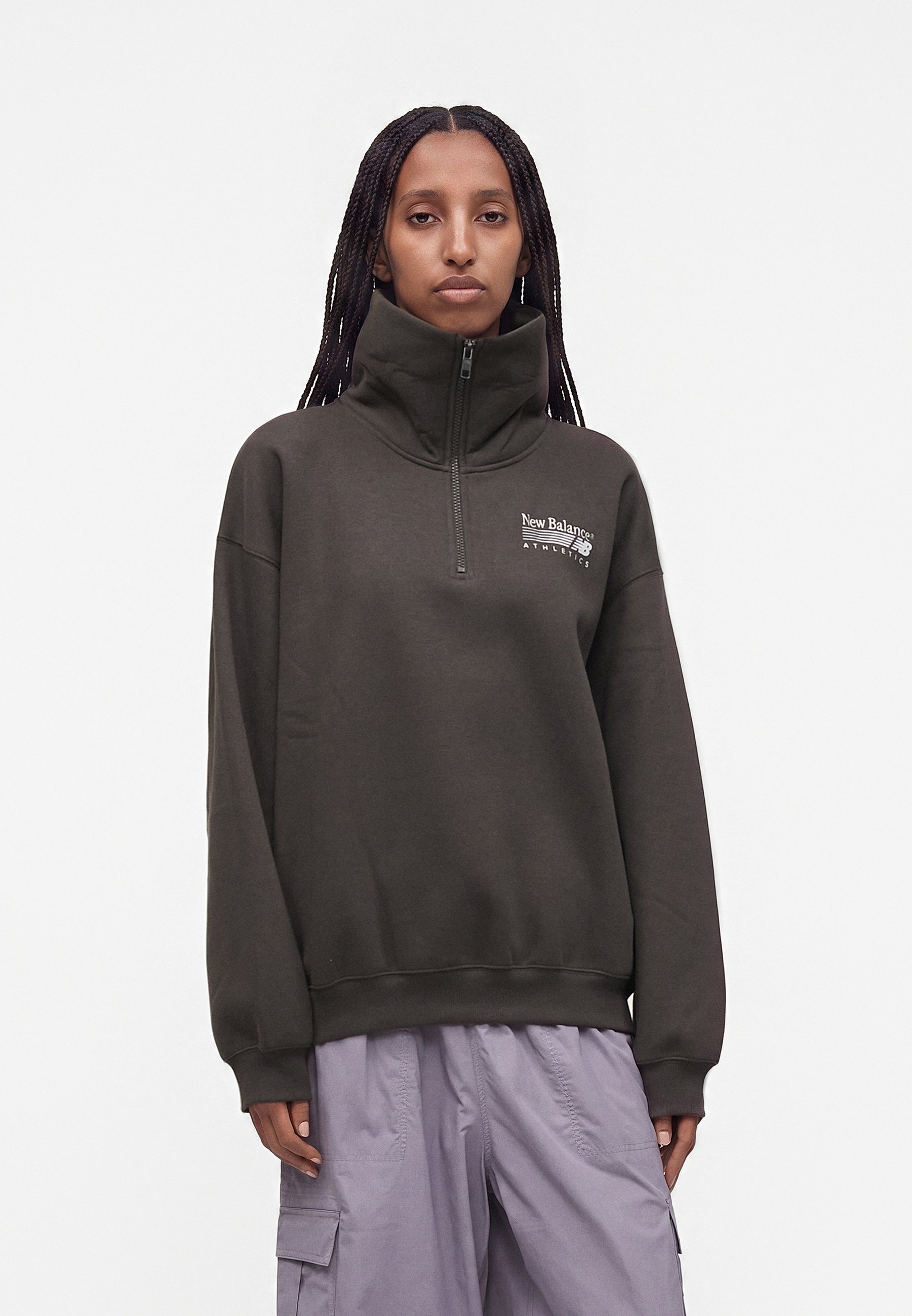 Mujer con el cabello trenzado, vistiendo un jersey New Balance gris oscuro con cremallera al cuarto y pantalones cargo de color lila claro, de pie frente a un fondo liso.