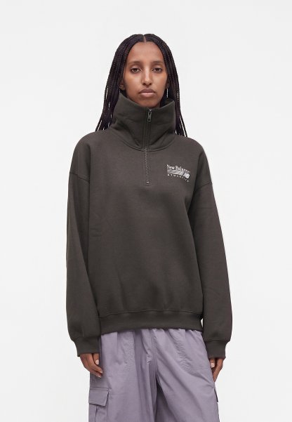 Mujer con el cabello trenzado, vistiendo un jersey New Balance gris oscuro con cremallera al cuarto y pantalones cargo de color lila claro, de pie frente a un fondo liso.