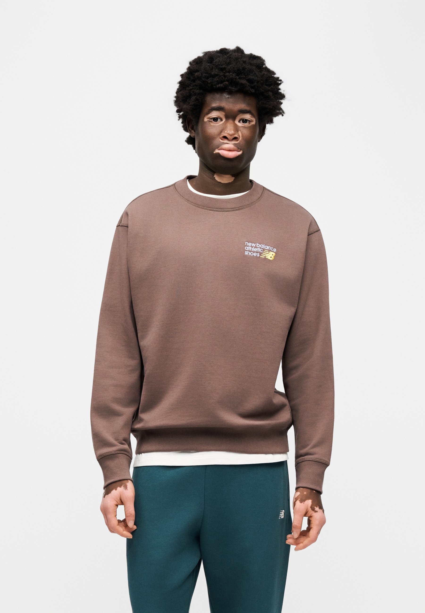Sudadera de algodón marrón con puños y cuello acanalados, con un pequeño logo de New Balance. Combinada con pantalones deportivos azul verdoso oscuro.