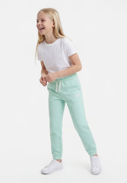 Niña joven riendo, vestida con una camiseta blanca, pantalones deportivos verde menta con cordón, y zapatillas blancas, de pie contra un fondo liso.