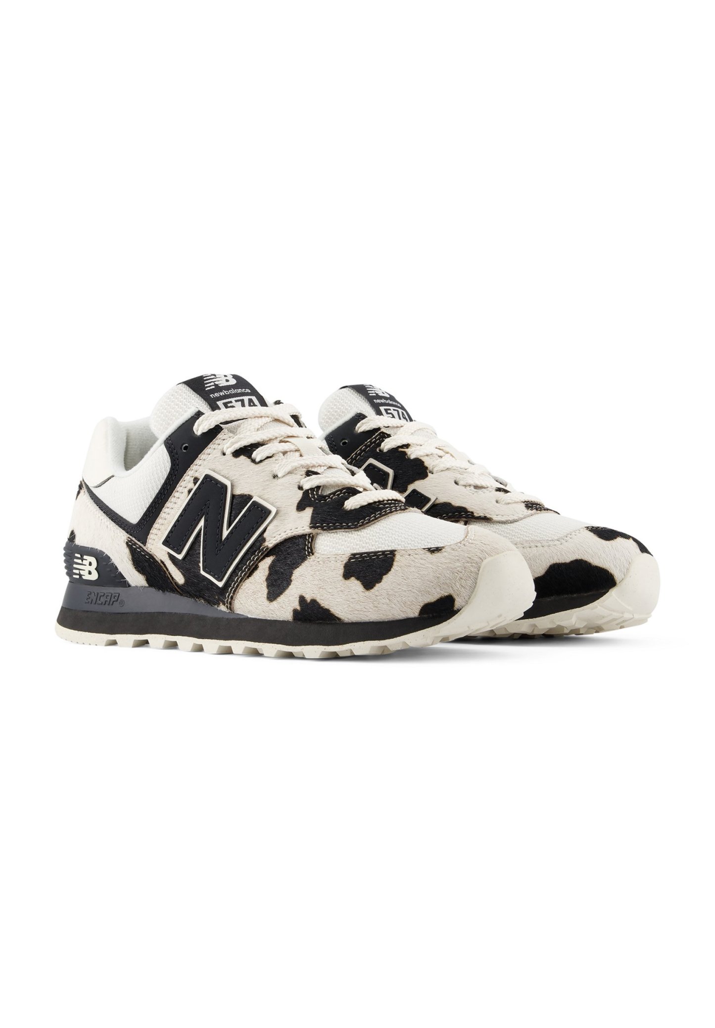 Par de zapatillas New Balance 574 con estampado de vaca en blanco y negro, logotipo negro y cordones blancos sobre fondo blanco.