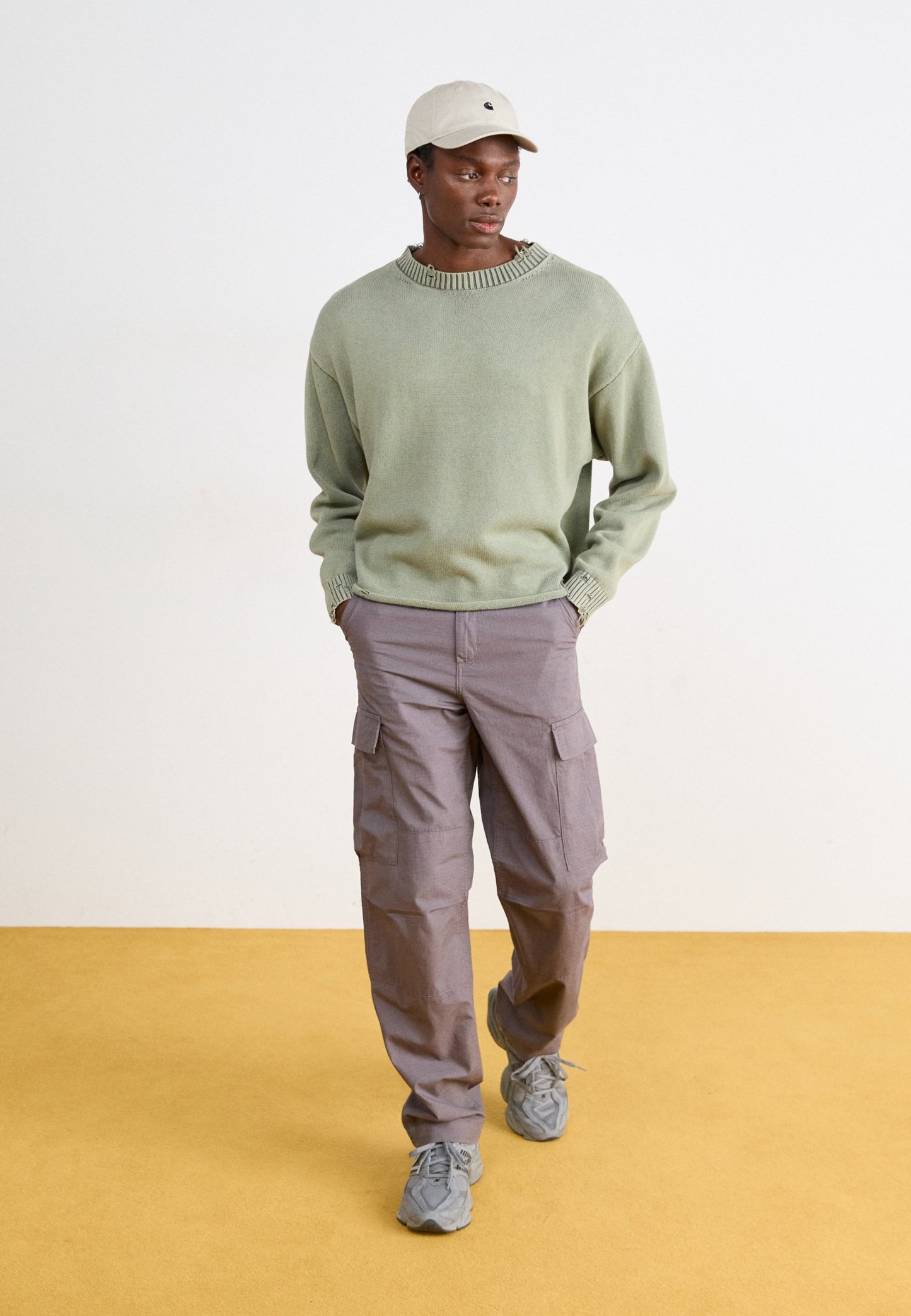 Hombre con suéter verde claro, gorra beige, pantalones cargo grises y zapatillas grises caminando sobre un suelo mostaza con fondo blanco.