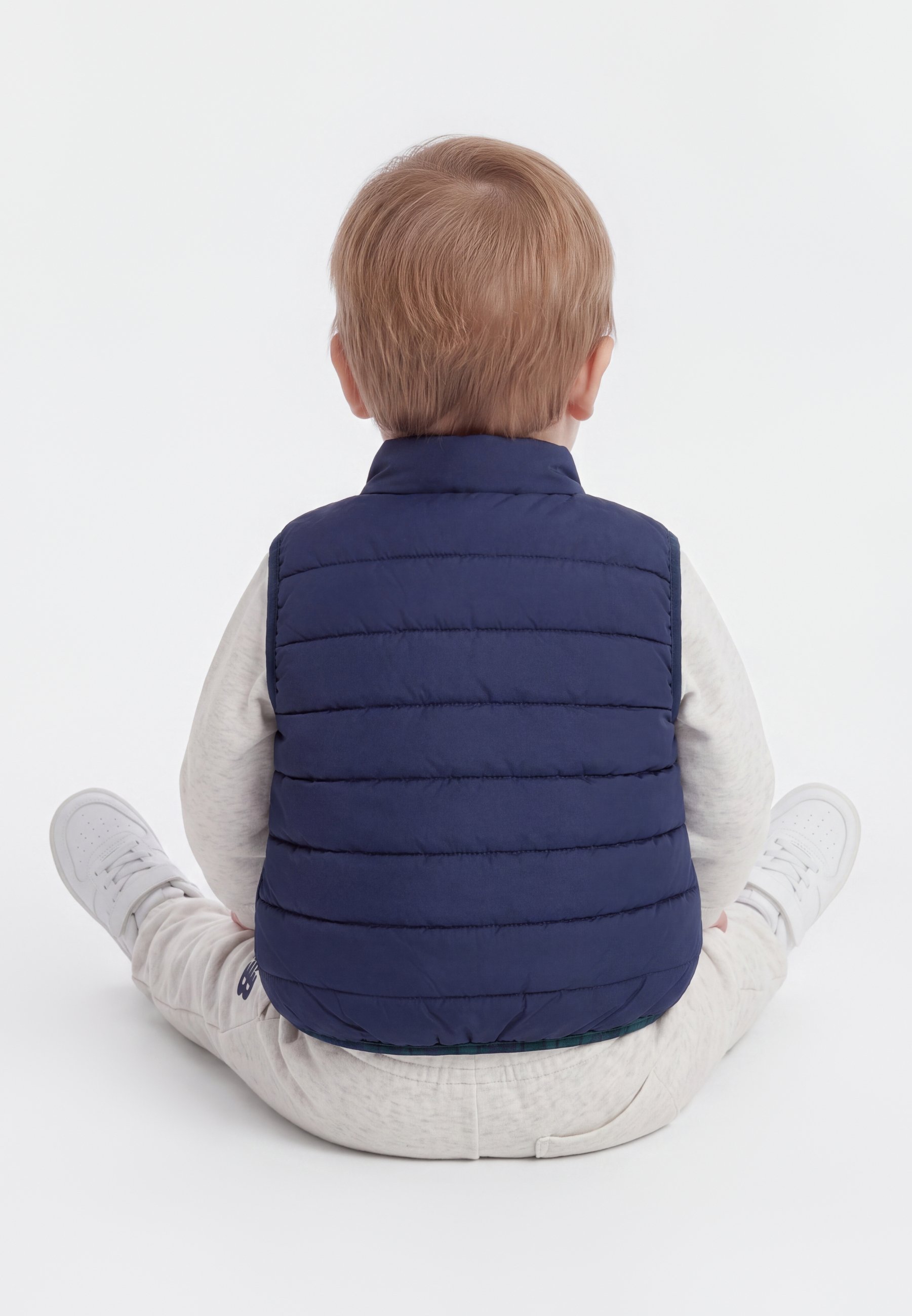 Niño pequeño con cabello castaño claro sentado con las piernas cruzadas, llevando un chaleco acolchado azul marino, pantalones de color claro y zapatillas blancas, de espaldas.