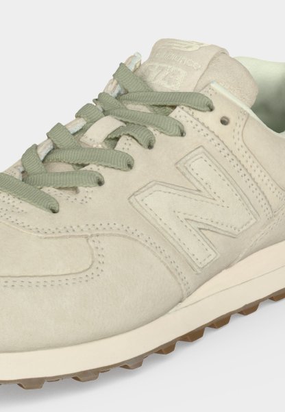 Zapatilla New Balance color beige claro con textura de ante, cordones de tela verdes, logo en relieve en el lateral y borde de suela de goma texturizada visible.