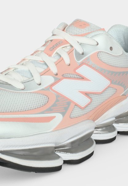 Zapatilla deportiva New Balance blanca y melocotón con tejido de malla, cierre de cordones, gran logotipo "N" y suela plateada segmentada para amortiguación.