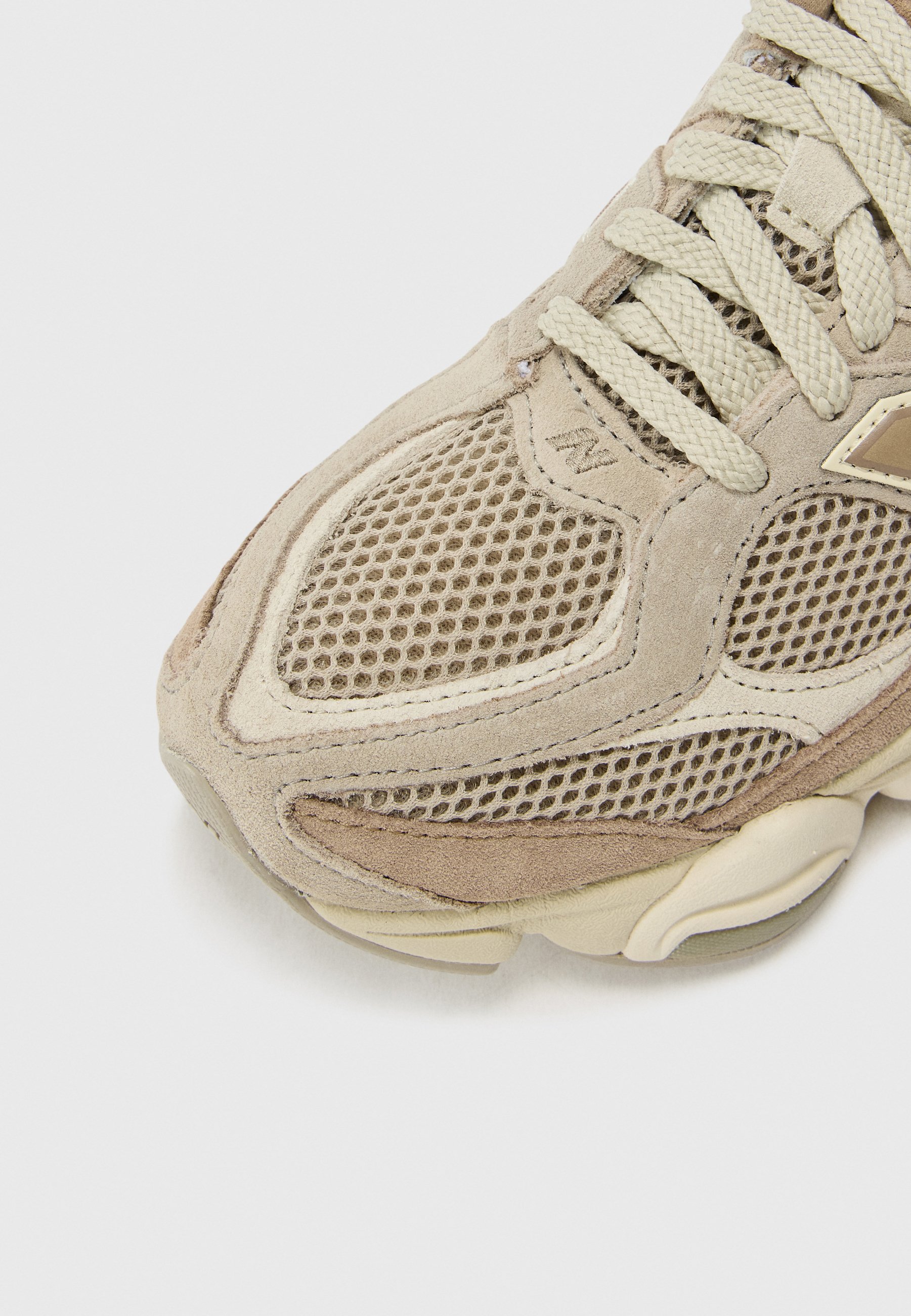Zapatilla deportiva beige que presenta materiales de ante y malla, detalles texturizados, puntera redondeada y diseño de suela de goma lisa. Cordonada con agujetas de color crema claro.
