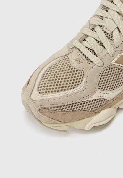 Zapatilla deportiva beige que presenta materiales de ante y malla, detalles texturizados, puntera redondeada y diseño de suela de goma lisa. Cordonada con agujetas de color crema claro.