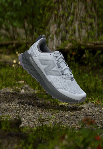 Zapatilla de running New Balance gris clara con suela acolchada flotando sobre un camino de grava en un entorno natural al aire libre.