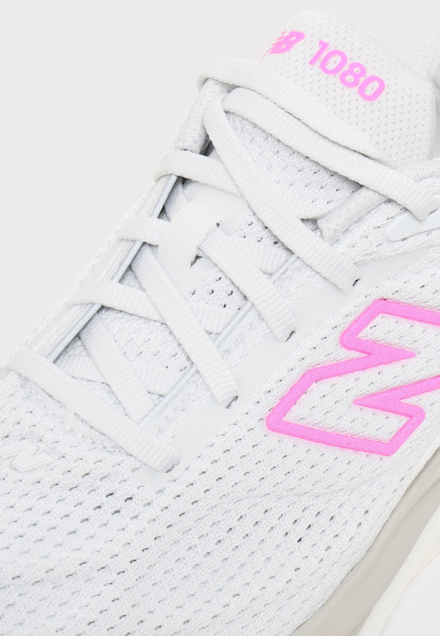 Primer plano de una zapatilla blanca New Balance 1080 con logo y marca de color rosa, con tejido de malla y cordones blancos cruzados.
