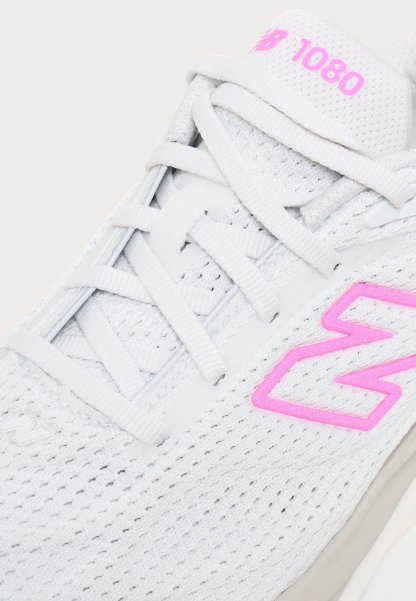 Primer plano de una zapatilla blanca New Balance 1080 con logo y marca de color rosa, con tejido de malla y cordones blancos cruzados.