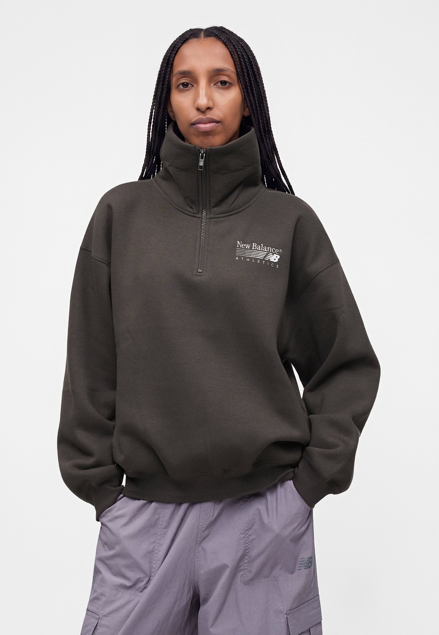 Persona con cabello largo trenzado que lleva una sudadera New Balance de color verde oscuro con media cremallera y pantalones cargo lila claro, de pie con las manos en los bolsillos.
