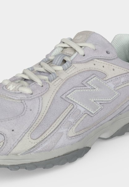 Zapatilla deportiva de ante gris claro con cordones blancos, cuello acolchado y un gran logo "N" cosido en el lateral, sobre una suela de goma gris.