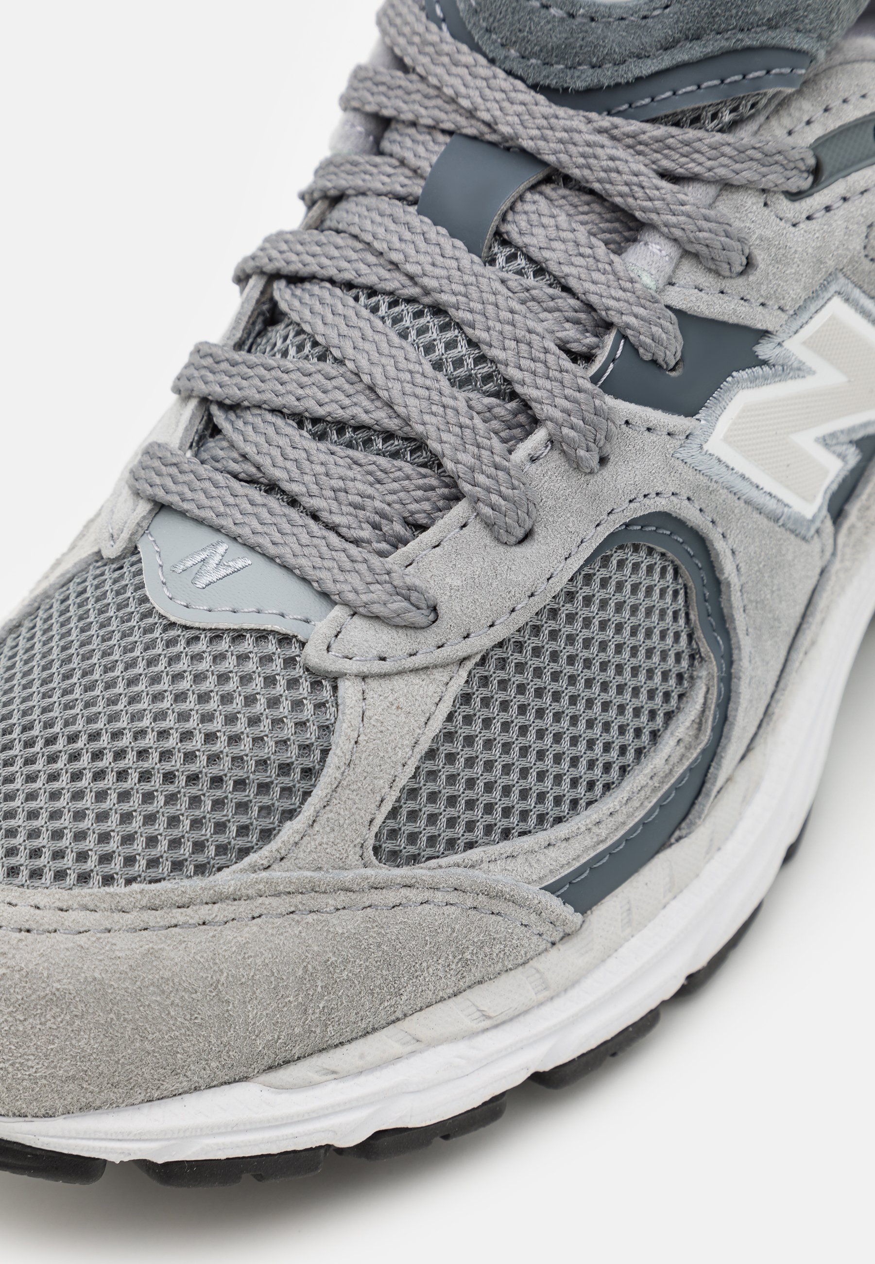 Zapatilla atlética gris que presenta una combinación de materiales de malla y gamuza, cordones texturizados y un logotipo de acento en blanco en el costado.
