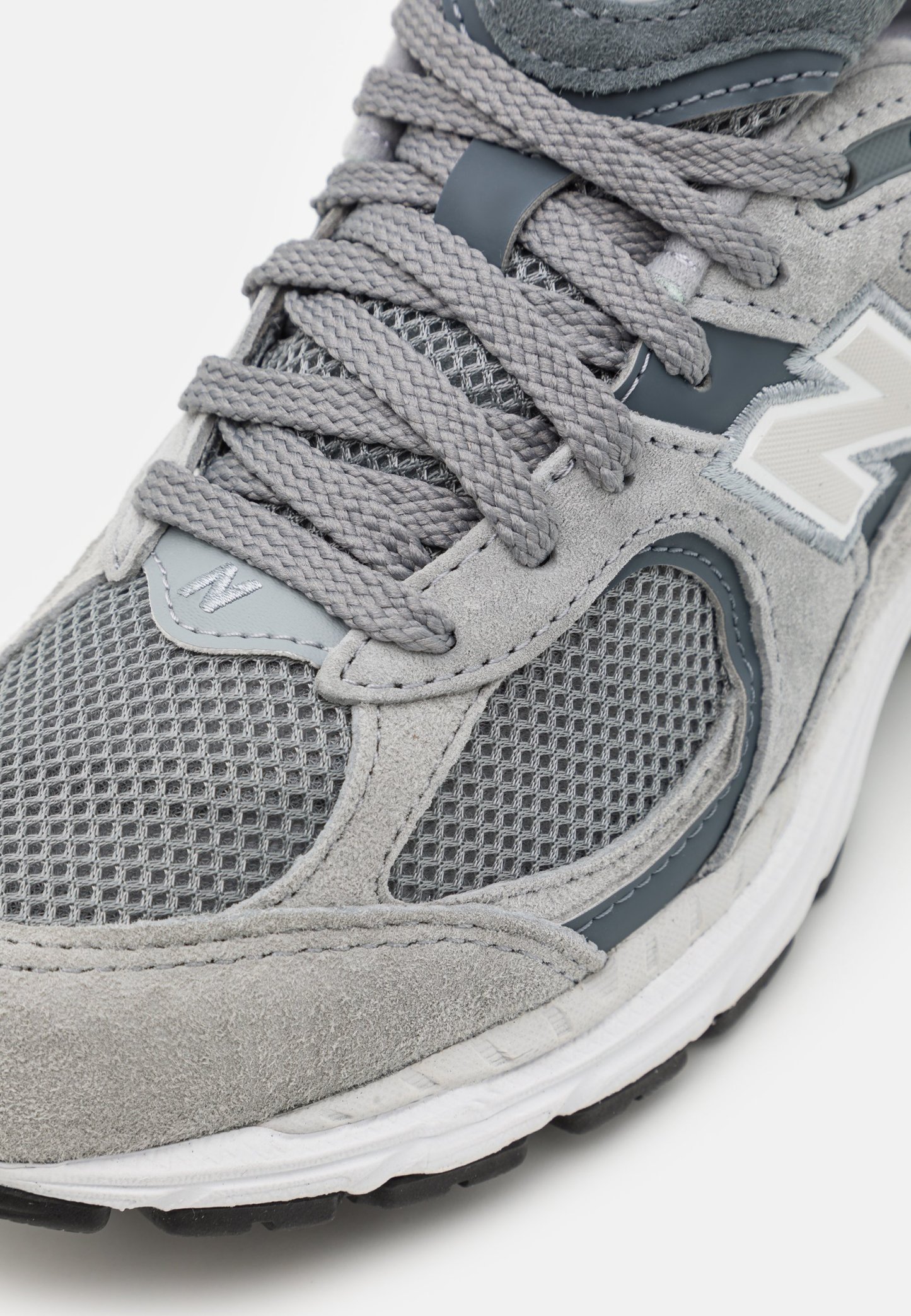 Zapatilla atlética gris que presenta una combinación de materiales de malla y gamuza, cordones texturizados y un logotipo de acento en blanco en el costado.