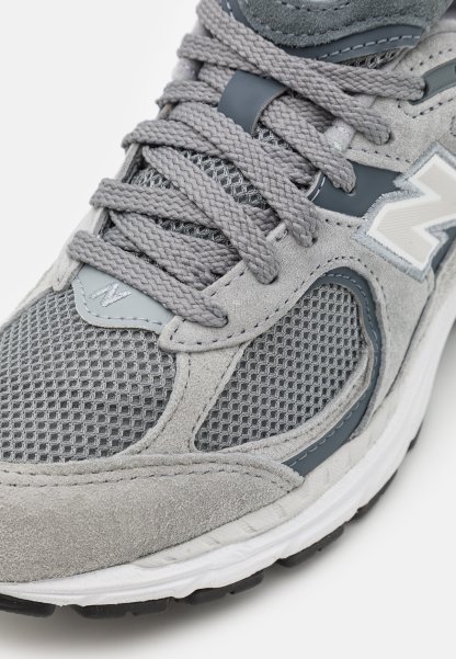 Zapatilla atlética gris que presenta una combinación de materiales de malla y gamuza, cordones texturizados y un logotipo de acento en blanco en el costado.