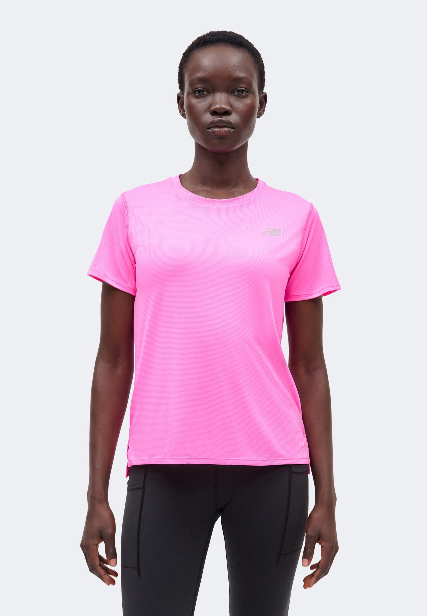 Mujer de pie con una camiseta deportiva de manga corta de color rosa brillante y pantalones negros ajustados, mirando al frente con expresión neutral.