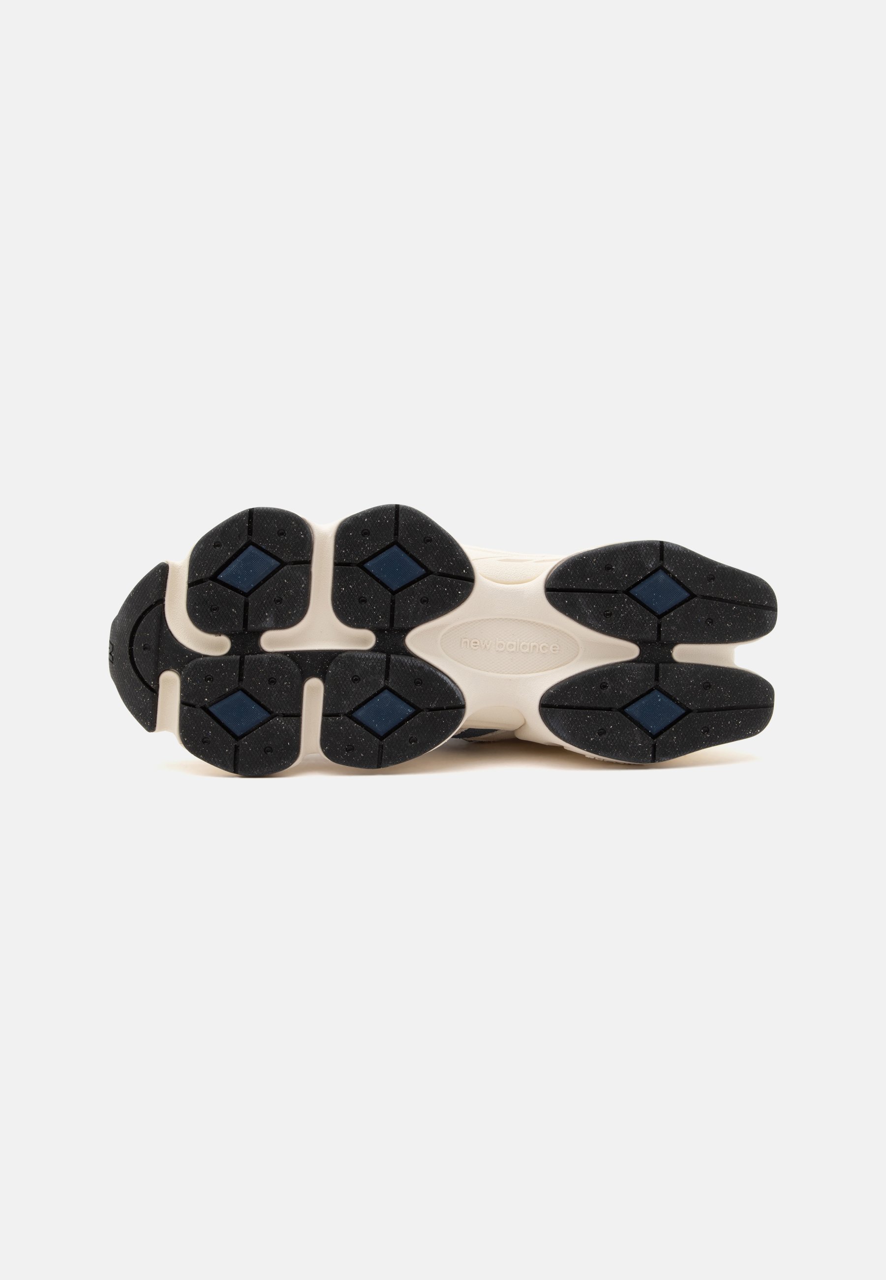Suela de una zapatilla New Balance que muestra almohadillas de goma negras y azules sobre una base color marfil contra un fondo liso y claro.