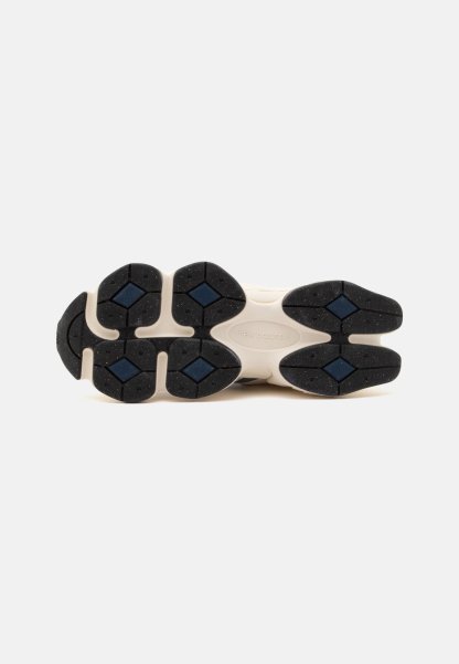 Suela de una zapatilla New Balance que muestra almohadillas de goma negras y azules sobre una base color marfil contra un fondo liso y claro.