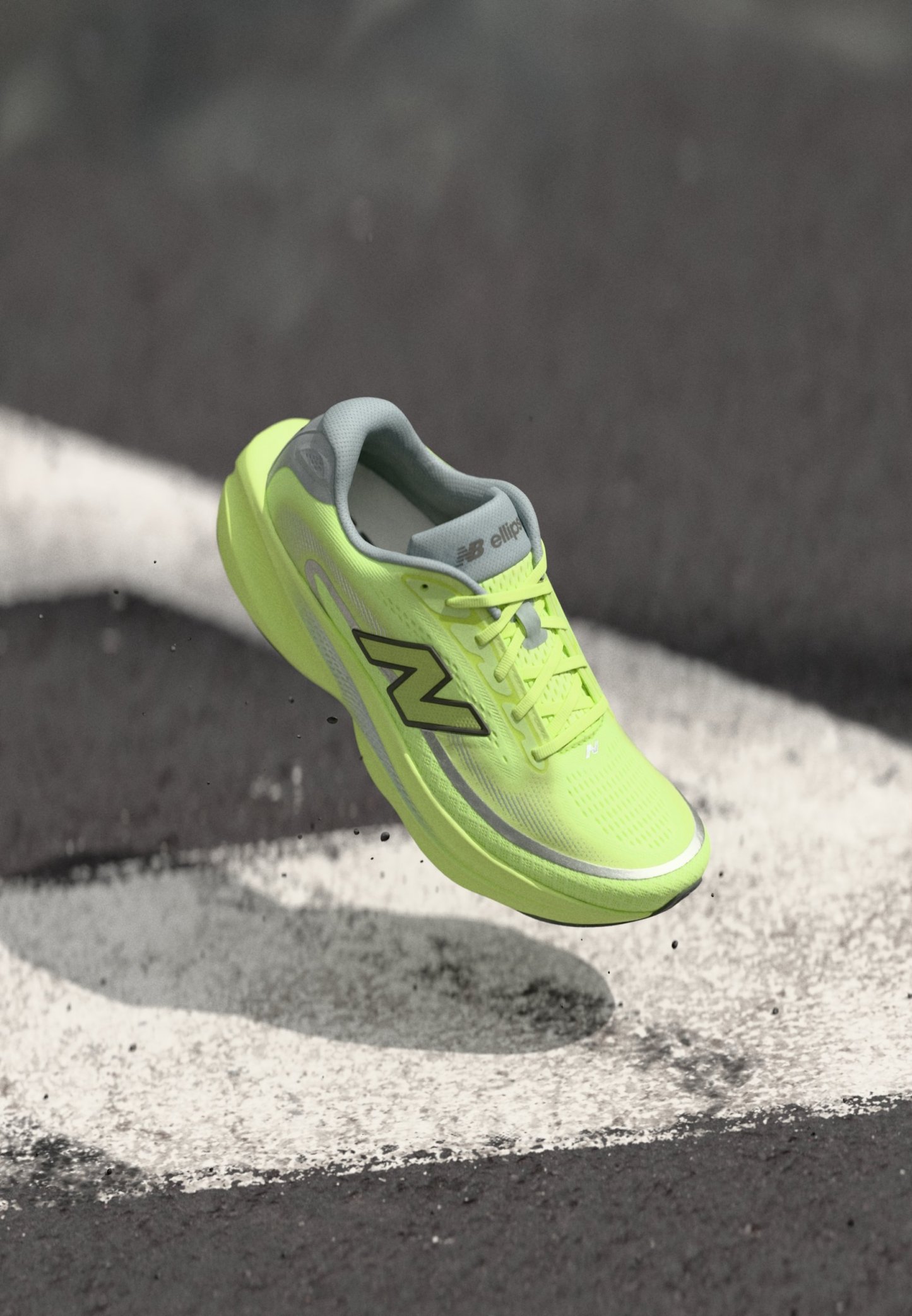 Zapatilla de running New Balance de neón amarillo en el aire sobre asfalto con marcas blancas pintadas y pequeños escombros debajo.