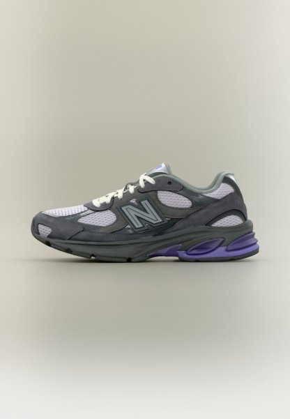 Zapatilla deportiva gris y blanca con acentos morados, paneles de malla y cordones blancos, vista de lado sobre un fondo neutro.