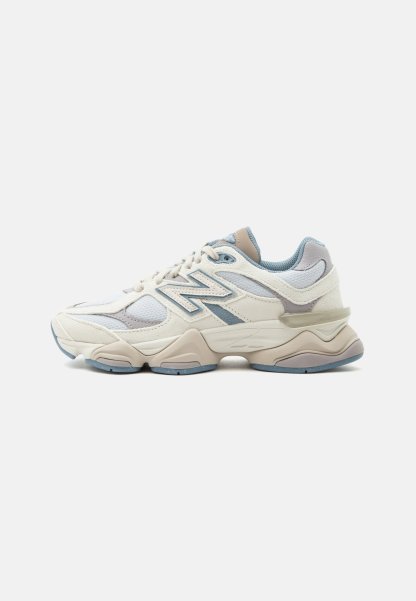 Zapatilla New Balance blanca y azul claro con parte superior de malla y ante, cierre de cordones y suela gruesa, mostrada de lado sobre fondo blanco.