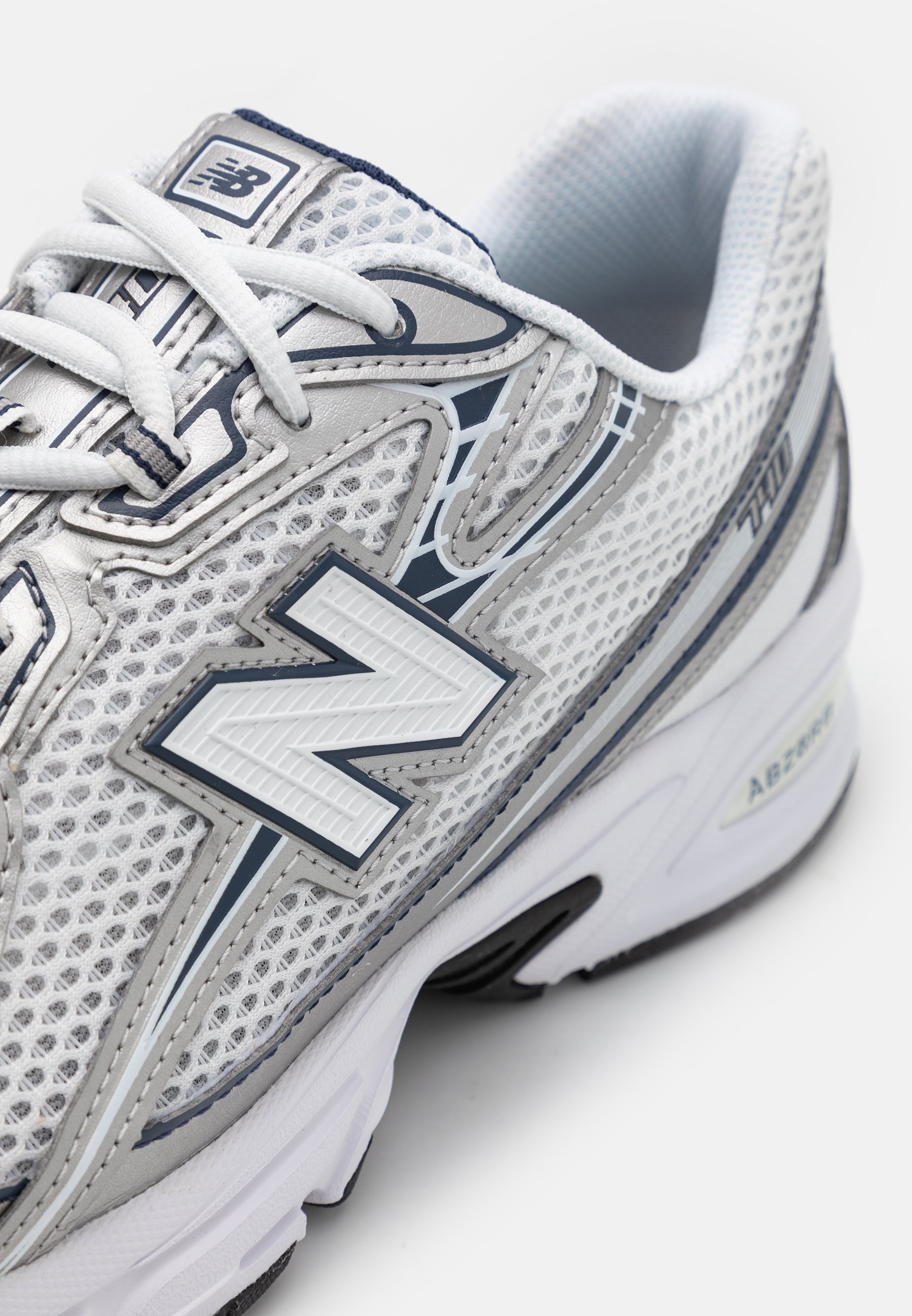 Zapatilla de running blanca de malla con acentos grises, que presenta un gran logo blanco con una "N", diseño con cordones y suela acolchada para mayor soporte.