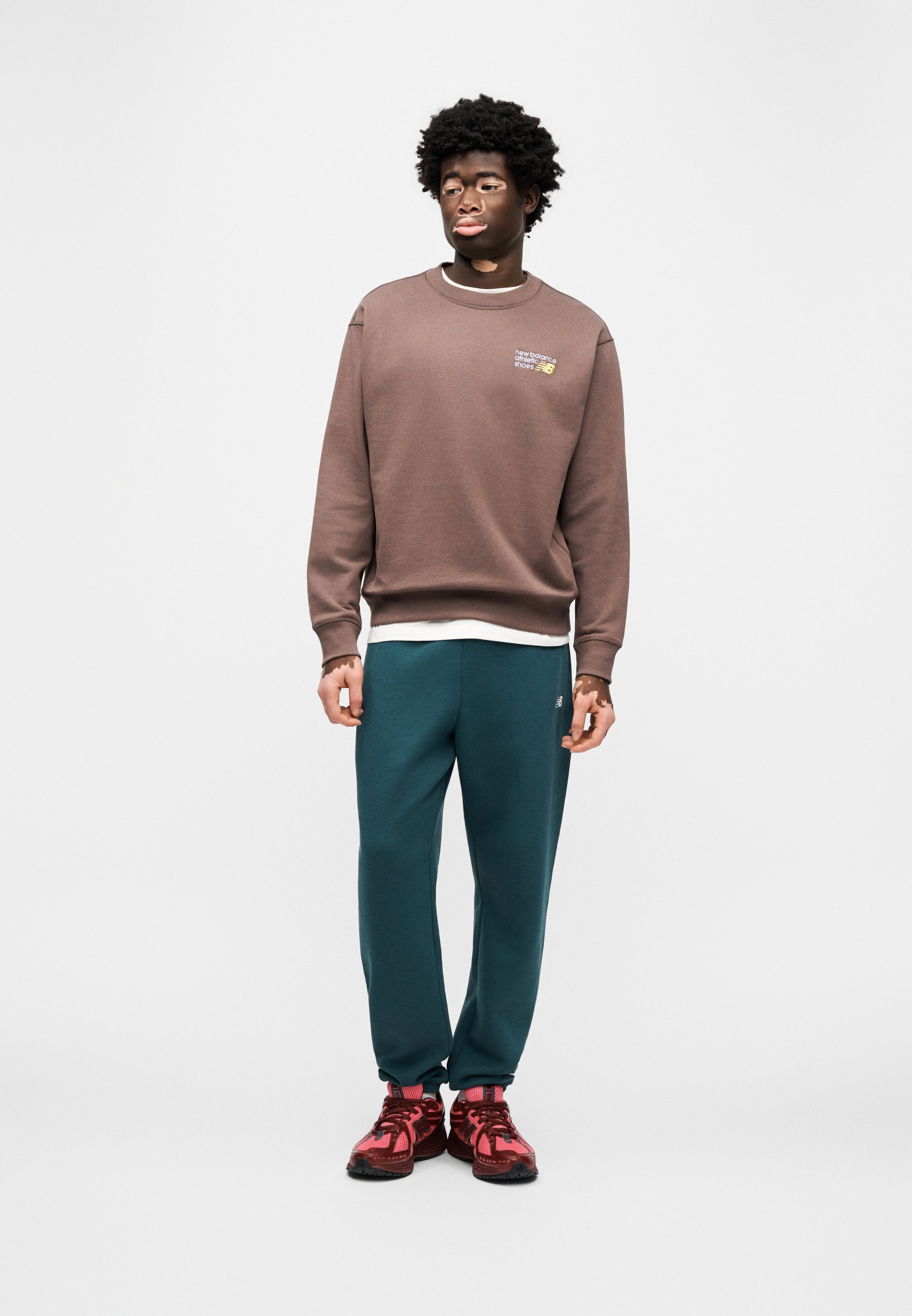 Sudadera marrón con cuello redondo y pequeño gráfico, pantalones deportivos verdes, zapatillas deportivas rojas con suelas negras, modelo de pie sobre un fondo liso.