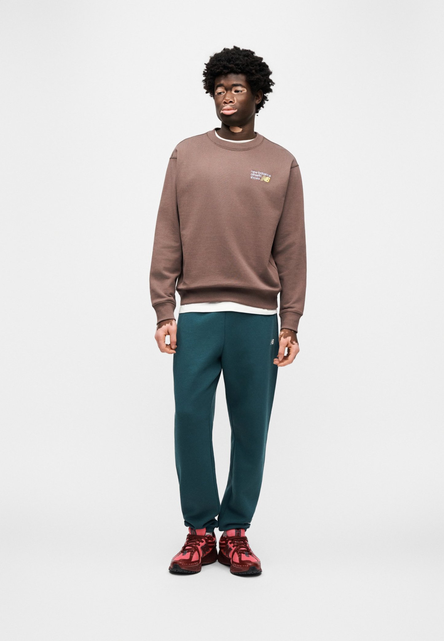 Sudadera marrón con cuello redondo y pequeño gráfico, pantalones deportivos verdes, zapatillas deportivas rojas con suelas negras, modelo de pie sobre un fondo liso.