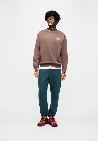 Sudadera marrón con cuello redondo y pequeño gráfico, pantalones deportivos verdes, zapatillas deportivas rojas con suelas negras, modelo de pie sobre un fondo liso.