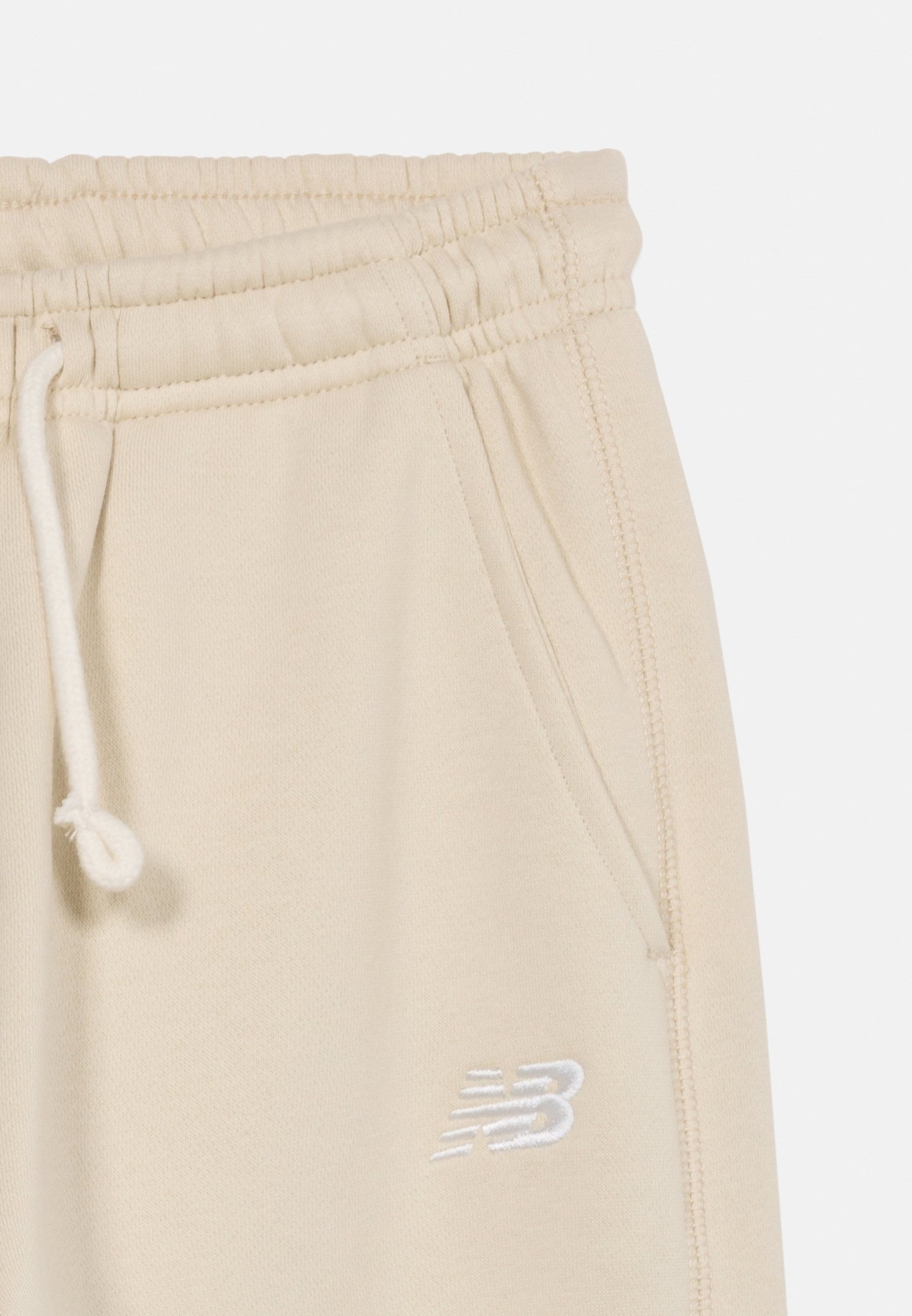Pantalones deportivos beige con cinturilla elástica, cordón blanco, bolsillo lateral y logo de New Balance bordado en blanco en el muslo.