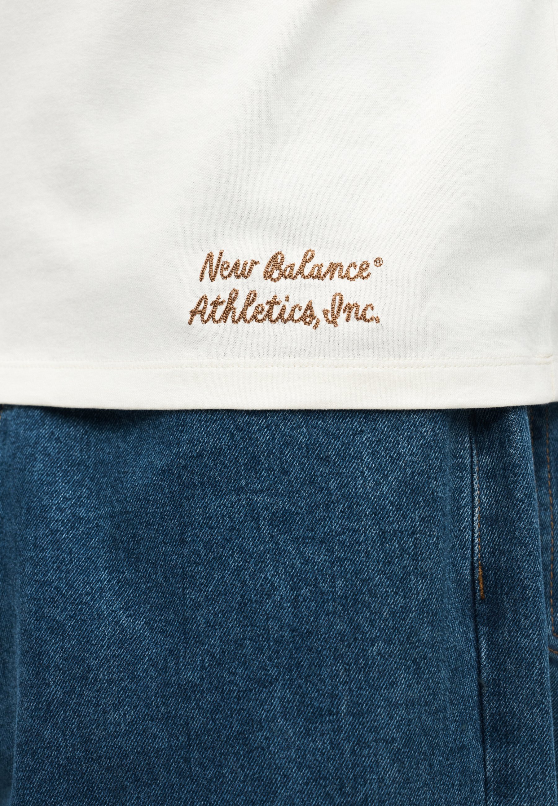 Dobladillo de camisa blanca con bordado marrón "New Balance Athletics, Inc." sobre pantalones vaqueros azules de denim.
